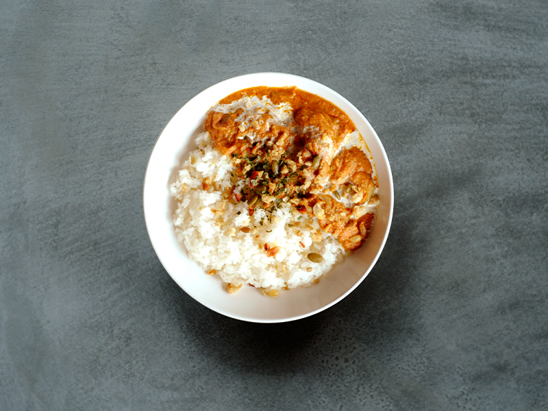 白いお皿に盛られたバターチキンカレー。とろみのある赤茶色のルーの中に、やわらかそうな鶏肉がごろっと入っている。横には白ごはん