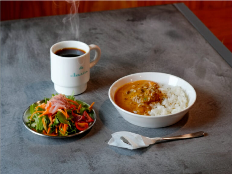 湯気の立つパイカカレーのプレートとホットコーヒー、サラダがモールテックスのテーブルに並ぶランチシーン。奥には木のカウンターが映り、温かく落ち着いた雰囲気のカフェの一角。