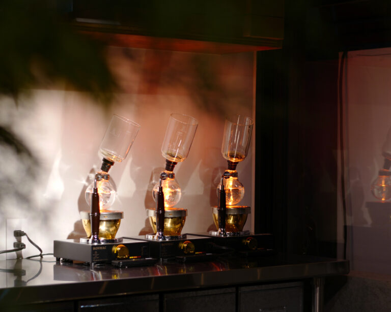 カフェのカウンターに並ぶサイフォン器具。温かな光に照らされ、ガラスフラスコが静かに抽出を行っている様子