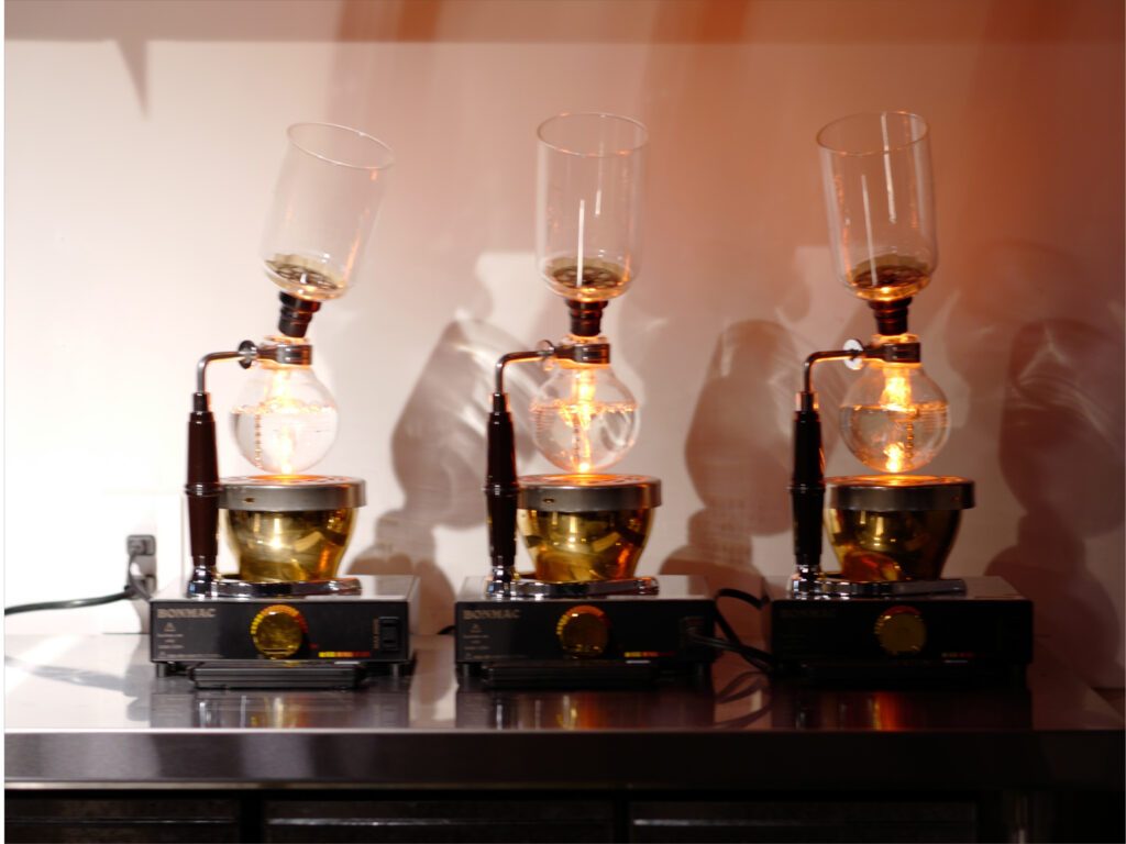 夜営業中の店内で、サイフォンでコーヒーを抽出している様子。ガラスボールに温かな光が映り込み、落ち着いた雰囲気の夜カフェ空間が写っている。