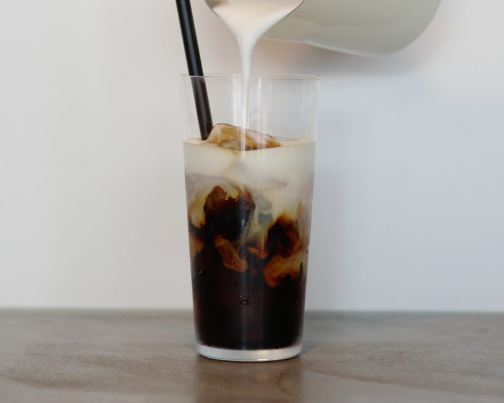 アイスカフェオレにミルクを注いでいる瞬間の写真。氷が入ったグラスにコーヒーとミルクが混ざり合い、層がゆっくり広がっている。ストローが入ったシンプルなグラスで、やさしい色合いと動きが印象的な一杯。