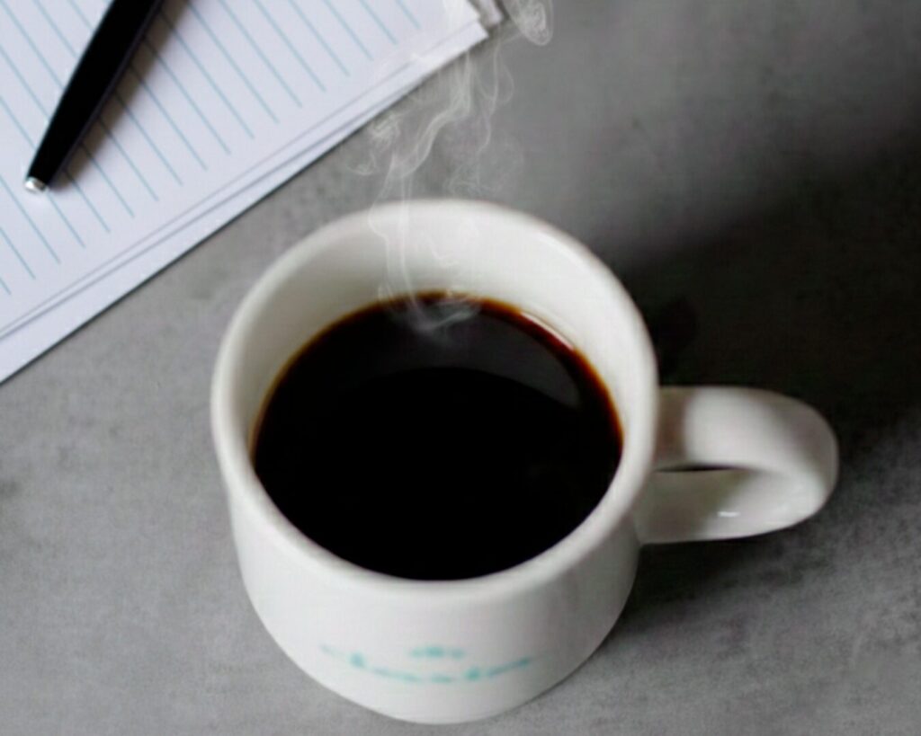 湯気の立つ黒いコーヒーが入った白いマグカップ。奥にはメモ帳とペンが置かれている。